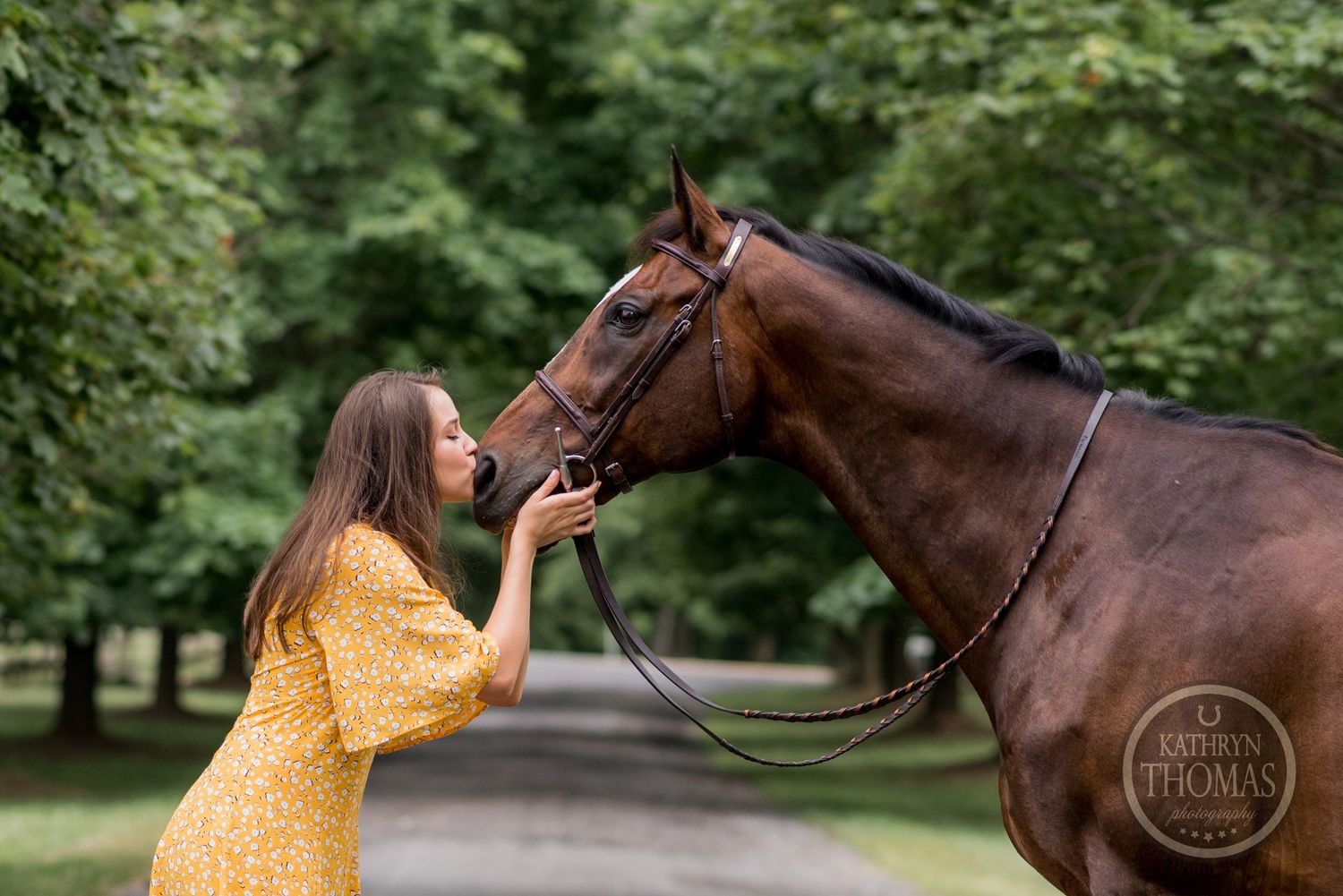 A tender moment between a bay horse and companion in a yellow dress on a wooded path.