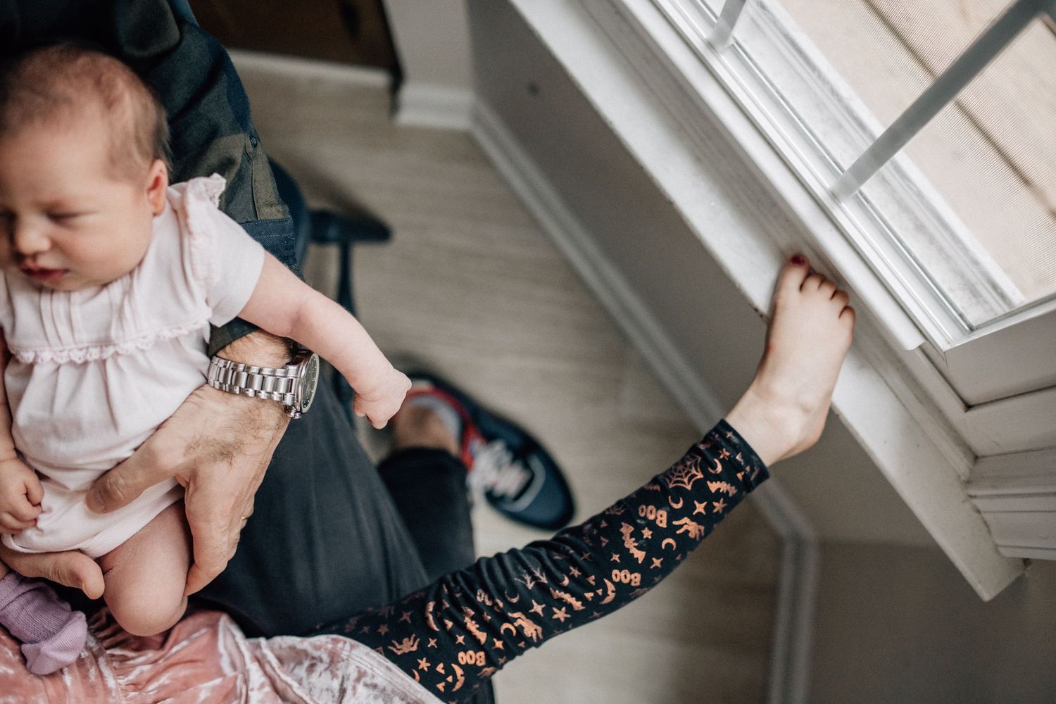 newborn being held by dad with a toddler leg on the window sill