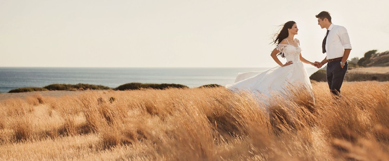 Couple in wedding attire walking through golden grass field by the ocean at sunset.