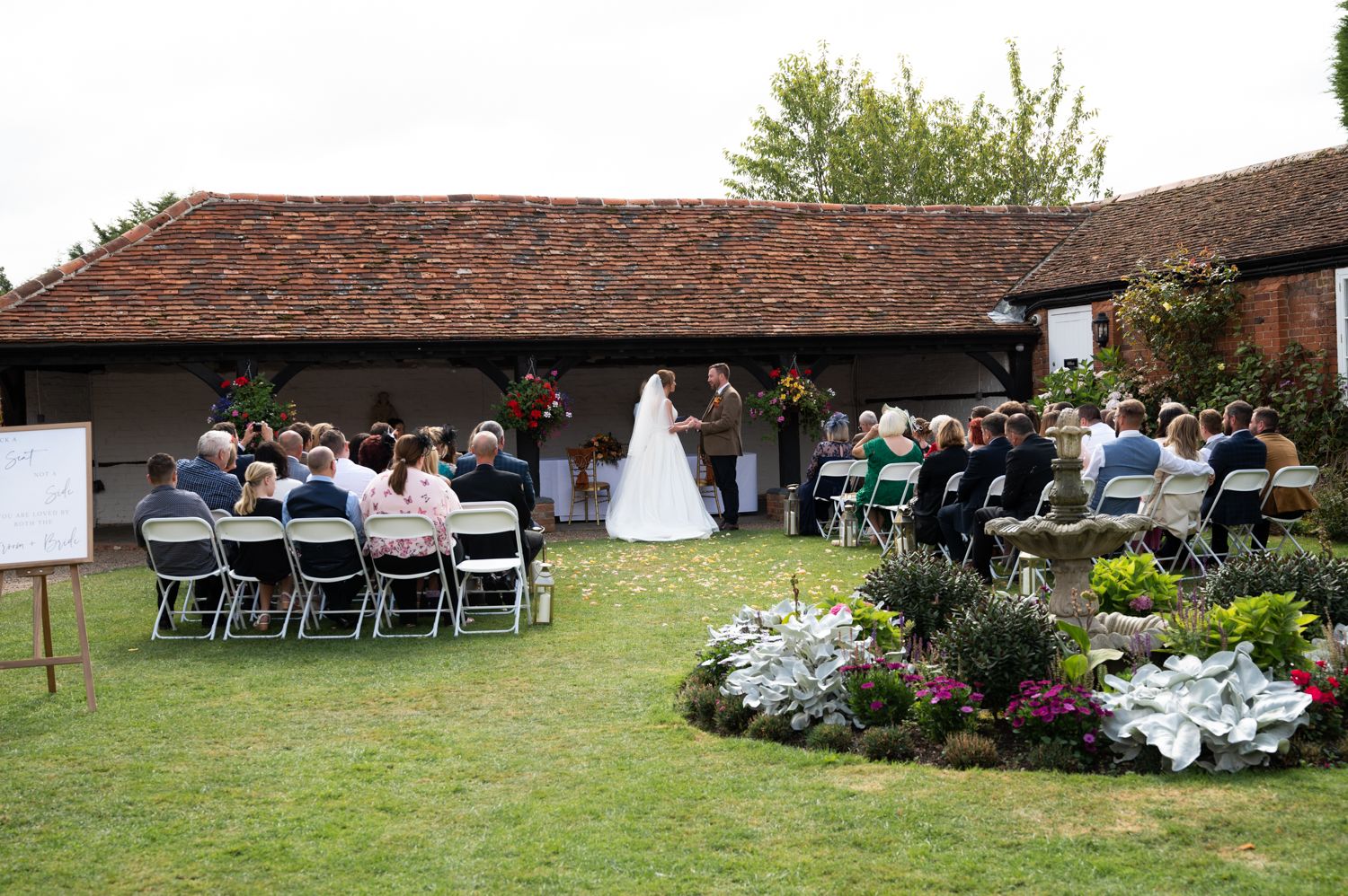 Outdoor wedding ceremony setup with white chairs and floral arrangements in garden setting.