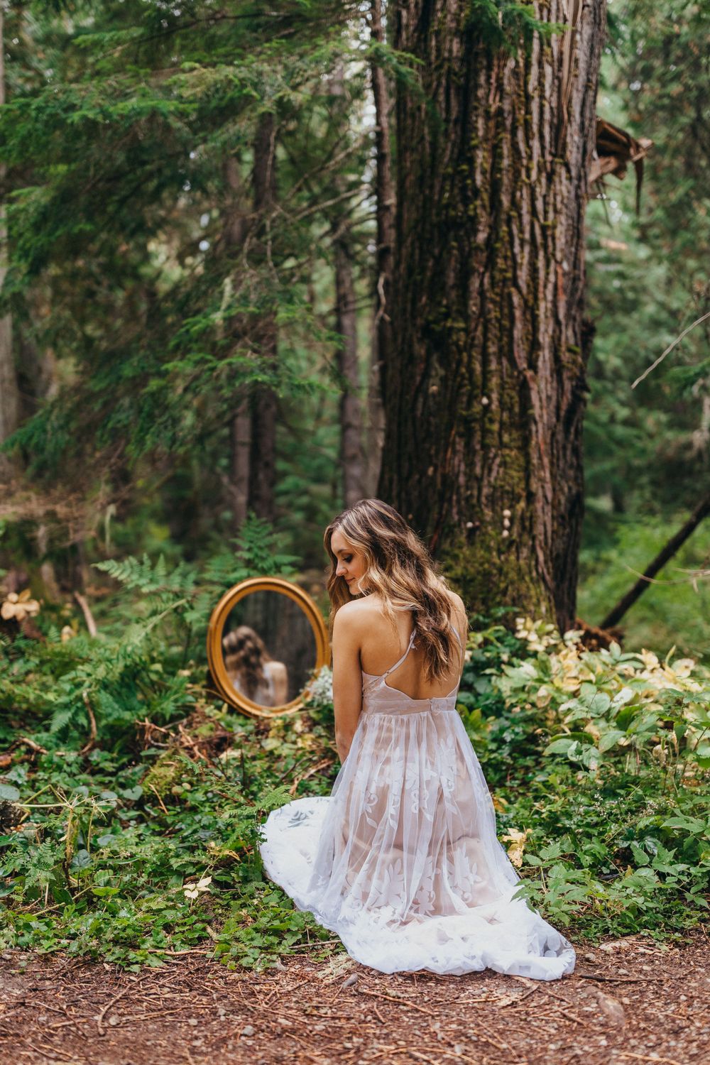 Woman in white lace dress kneeling with mirror in forest setting.