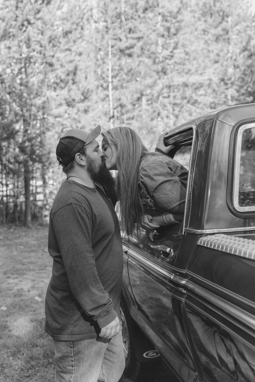 A couple shares a kiss by a vintage truck in a wooded setting.