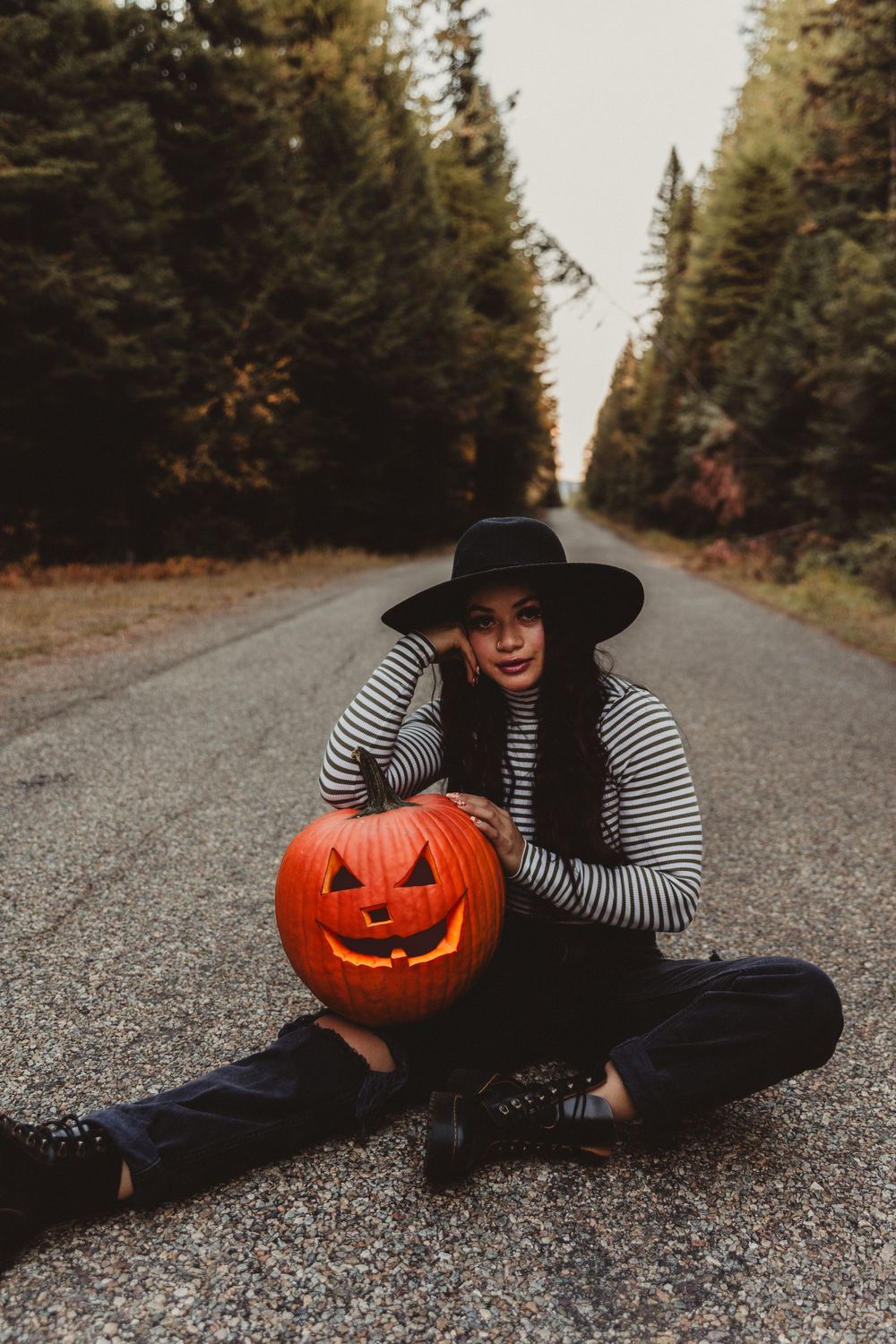 A person in a striped shirt and black hat poses with a carved jack-o-lantern on a forest road.