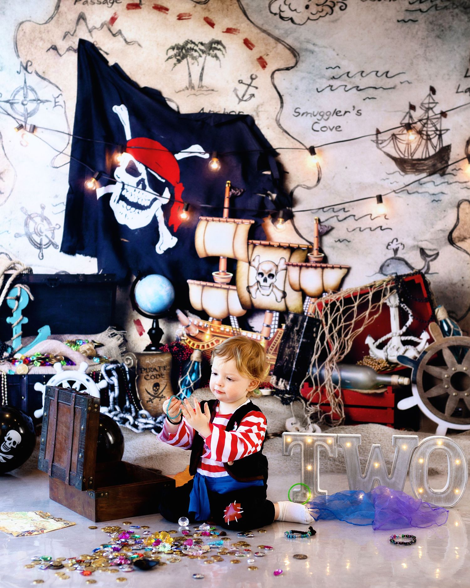 A young child in pirate costume plays with a treasure chest surrounded by nautical decor and a Jolly Roger flag.