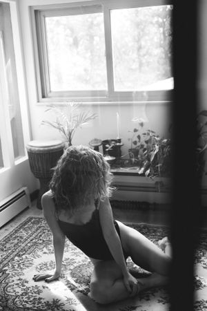 Person sitting by sunlit window with plants and rug in black and white composition.