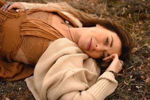 Person in cream sweater and brown clothing lying on autumn ground.