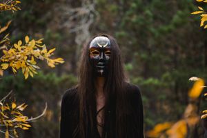 Person wearing black theatrical mask and dress poses among autumn foliage.