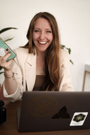 Woman in cream blazer smiles while working on laptop at desk with house plant in background.