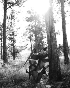 Black and white artistic photo of figure posed among tall pine trees in nature.