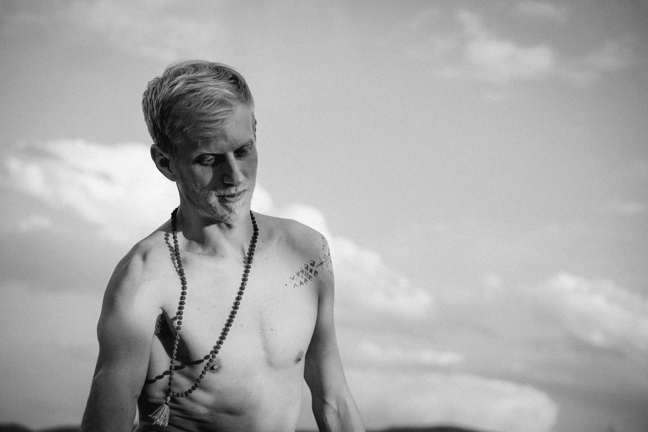Black and white portrait of a shirtless person wearing a necklace against cloudy sky.