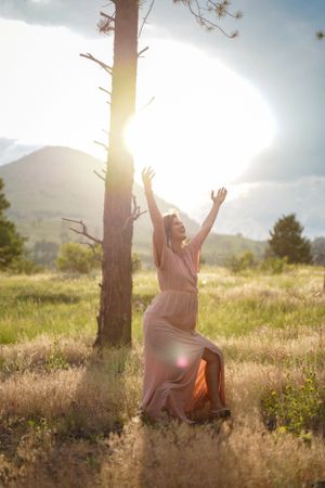 Figure in flowing dress dances in sunlit meadow with mountains and pine tree in background.