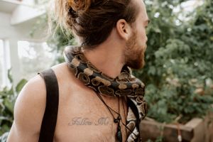 Close up of shoulder showing text tattoo and patterned scarf against green foliage.