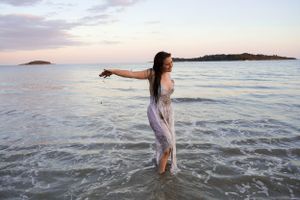 Someone in a flowing dress stands in shallow ocean water at sunset with arms outstretched.