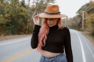 Person in a black crop top and brown hat stands on an empty rural road with pink hair.