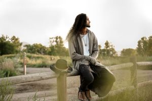 A casual portrait of someone sitting on a fence post at sunset in casual attire.