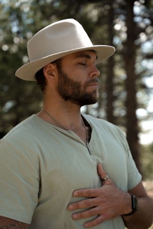 Person in white hat and t-shirt standing thoughtfully in nature with hand on chest.