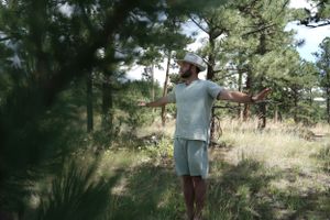 Figure standing in a forested area wearing casual clothing and looking into the distance.