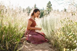 A person in a mauve dress sits peacefully among tall grass and wildflowers on a sunny day.
