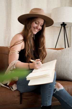Woman in brown hat writing in journal while relaxing on leather couch.