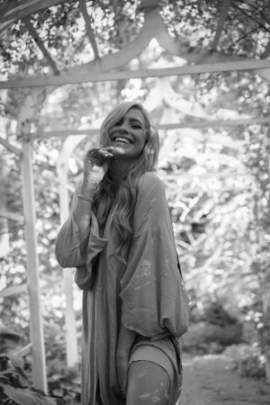 Black and white portrait of someone laughing joyfully under a pergola with dappled sunlight.