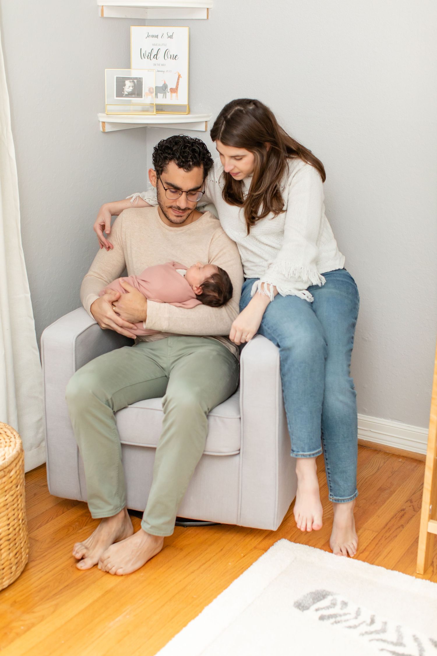 Couple sharing tender moment on grey armchair with sleeping newborn in nursery setting.