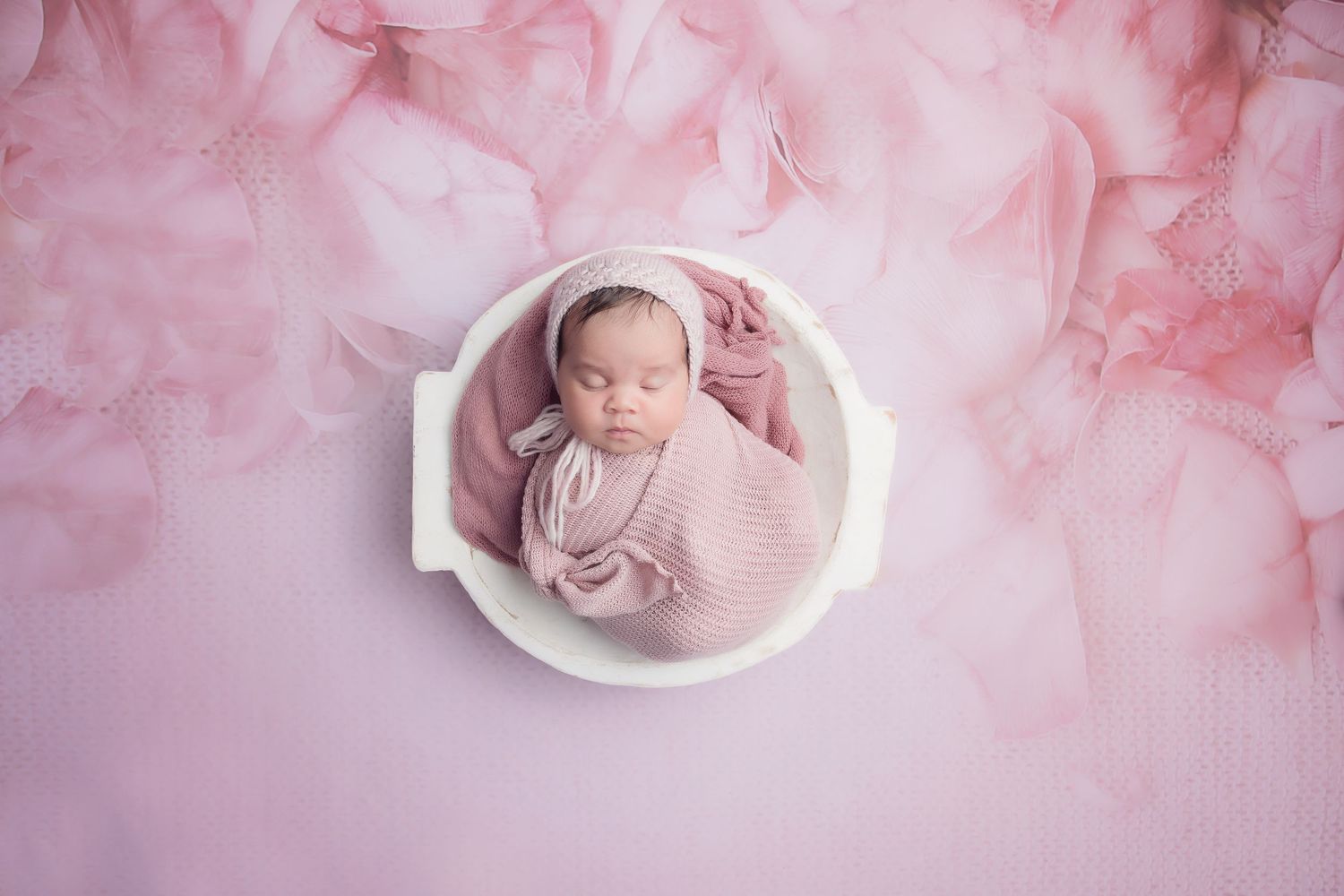 Sleeping newborn in pink bowl surrounded by soft pink tulle fabric.