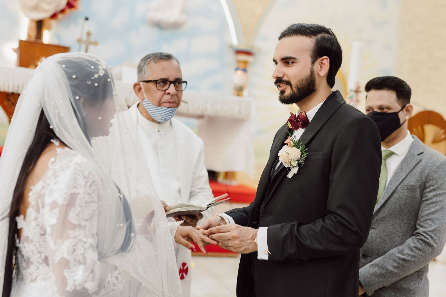 A wedding ceremony takes place in a church with a priest officiating as the couple exchanges rings.