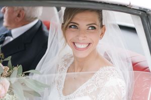 A radiant bride in white lace veil smiling brightly inside a car.