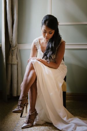 Elegant bride in a white gown and studded heels sits gracefully by a window in a vintage-styled room.