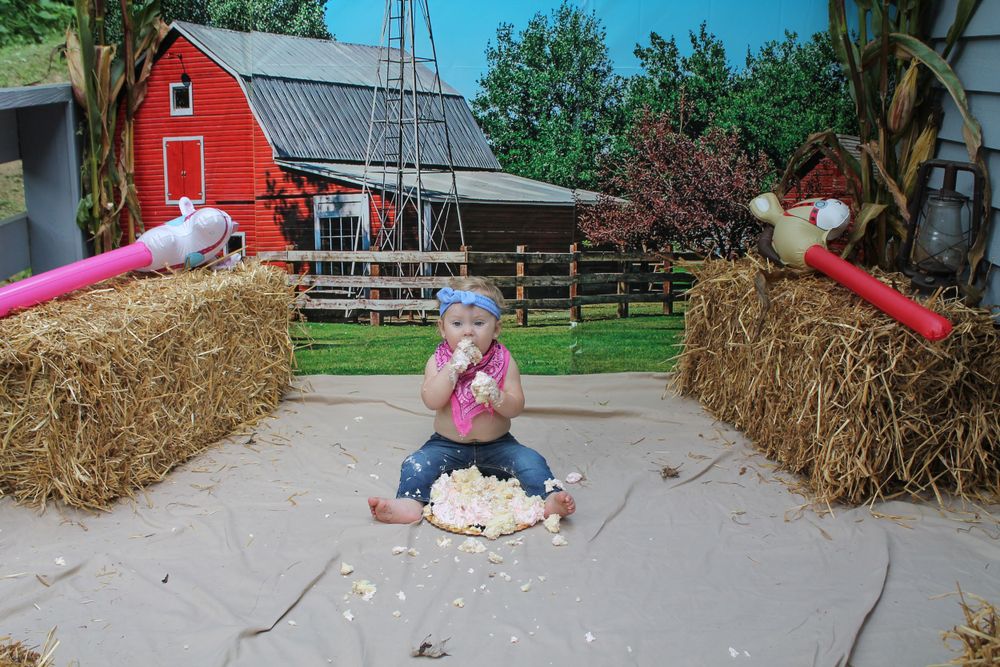 Small child sits between hay bales with red barn in background during smash cake photo session.