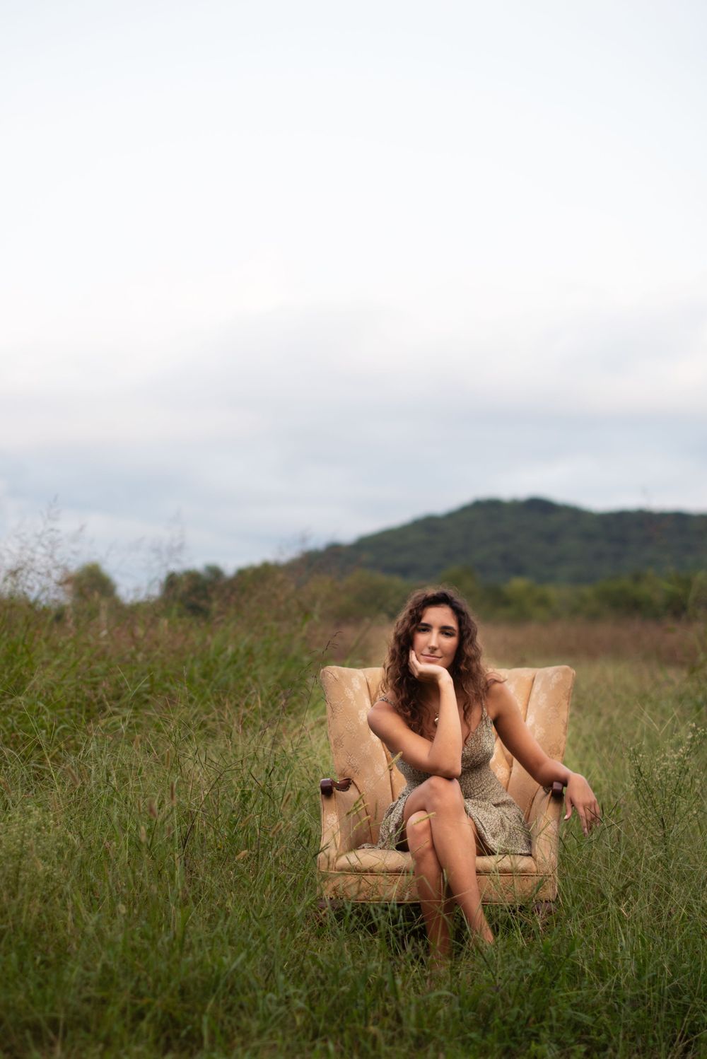A person sits thoughtfully in a vintage armchair placed in tall grass with mountains in the background.