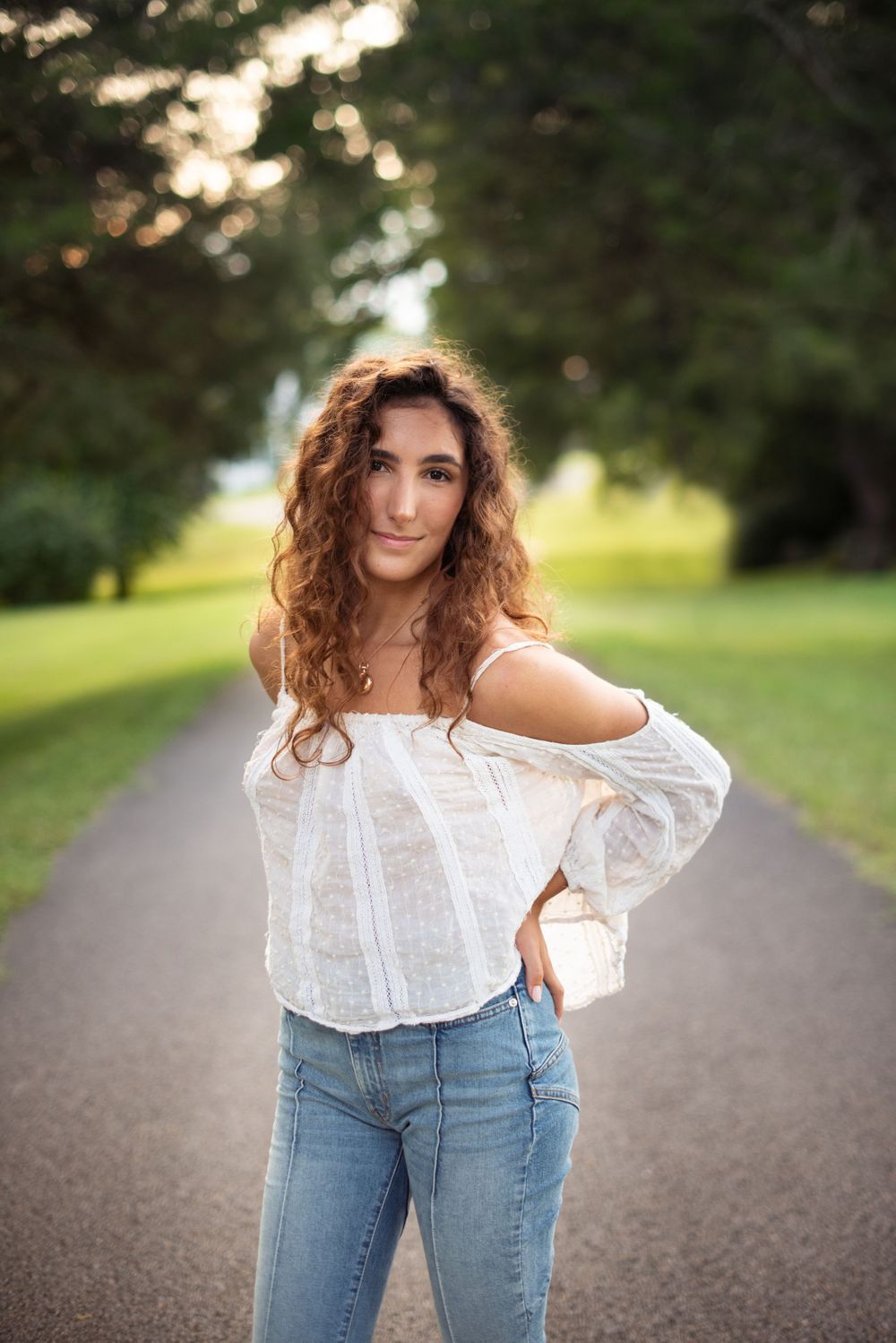 A person in a white blouse and jeans poses on a tree-lined path at sunset.