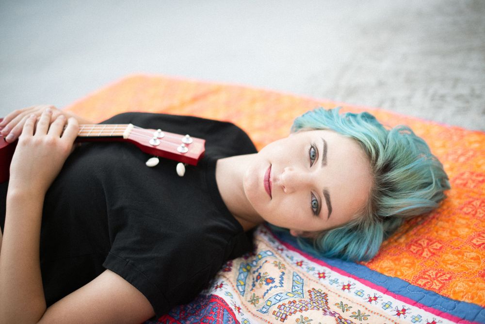 Person with teal hair lying on colorful blanket wearing black t-shirt.