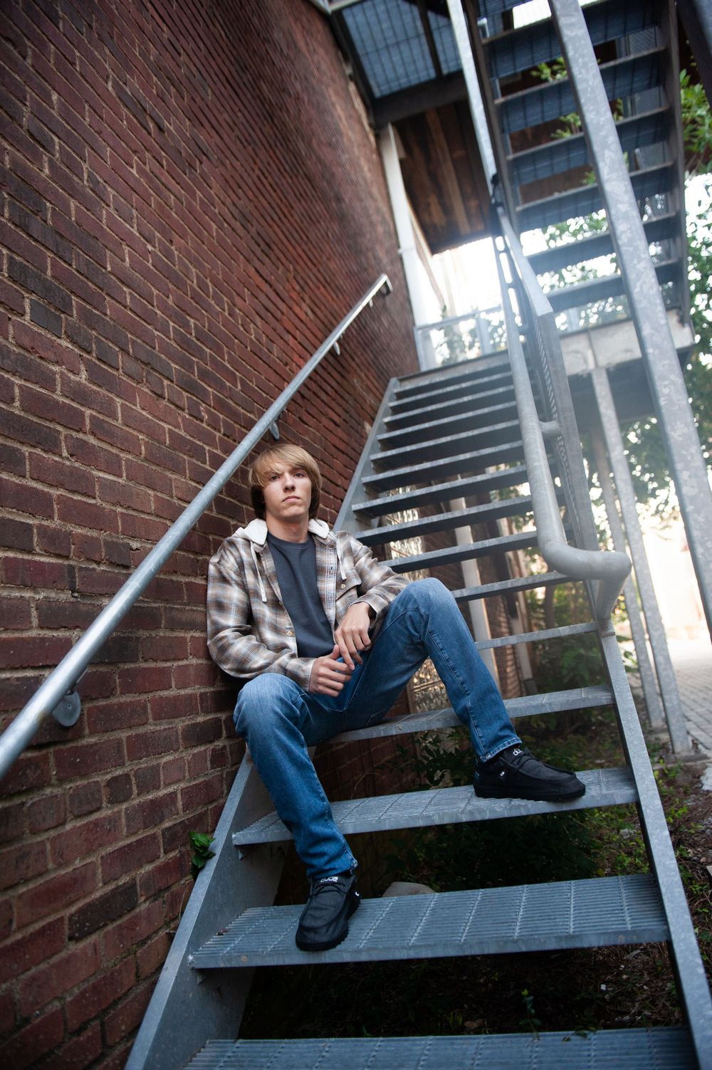 A person in a plaid shirt and jeans sits casually on metal stairs against a brick wall.