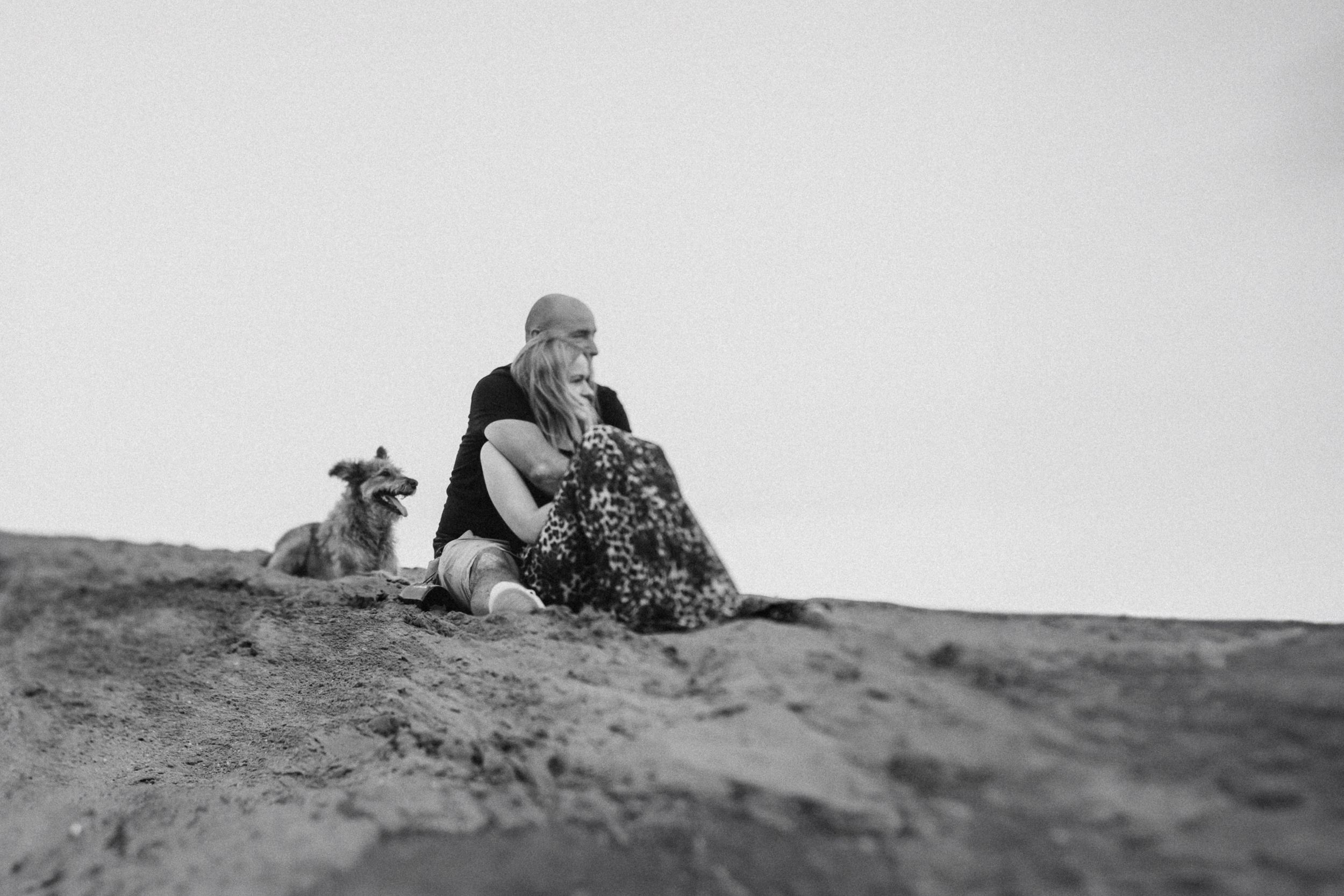 A person sits on sandy terrain with a dog beside them in a black and white photo.