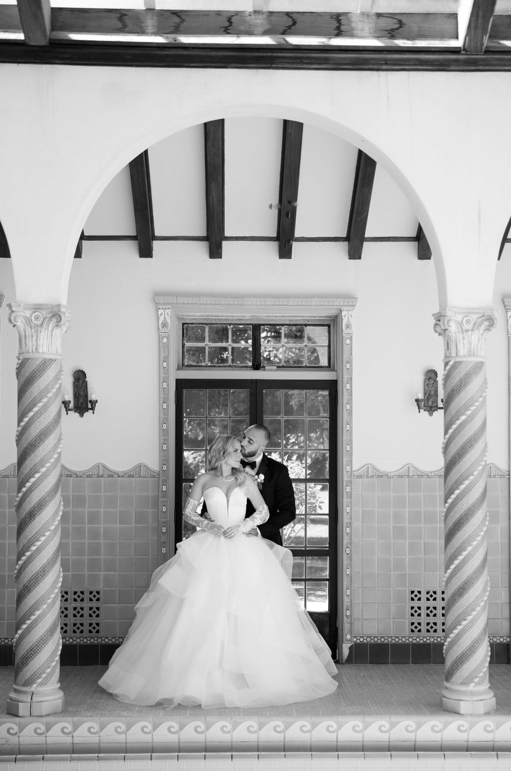 A romantic black and white wedding photo featuring an elegant couple posing under an ornate arched doorway.