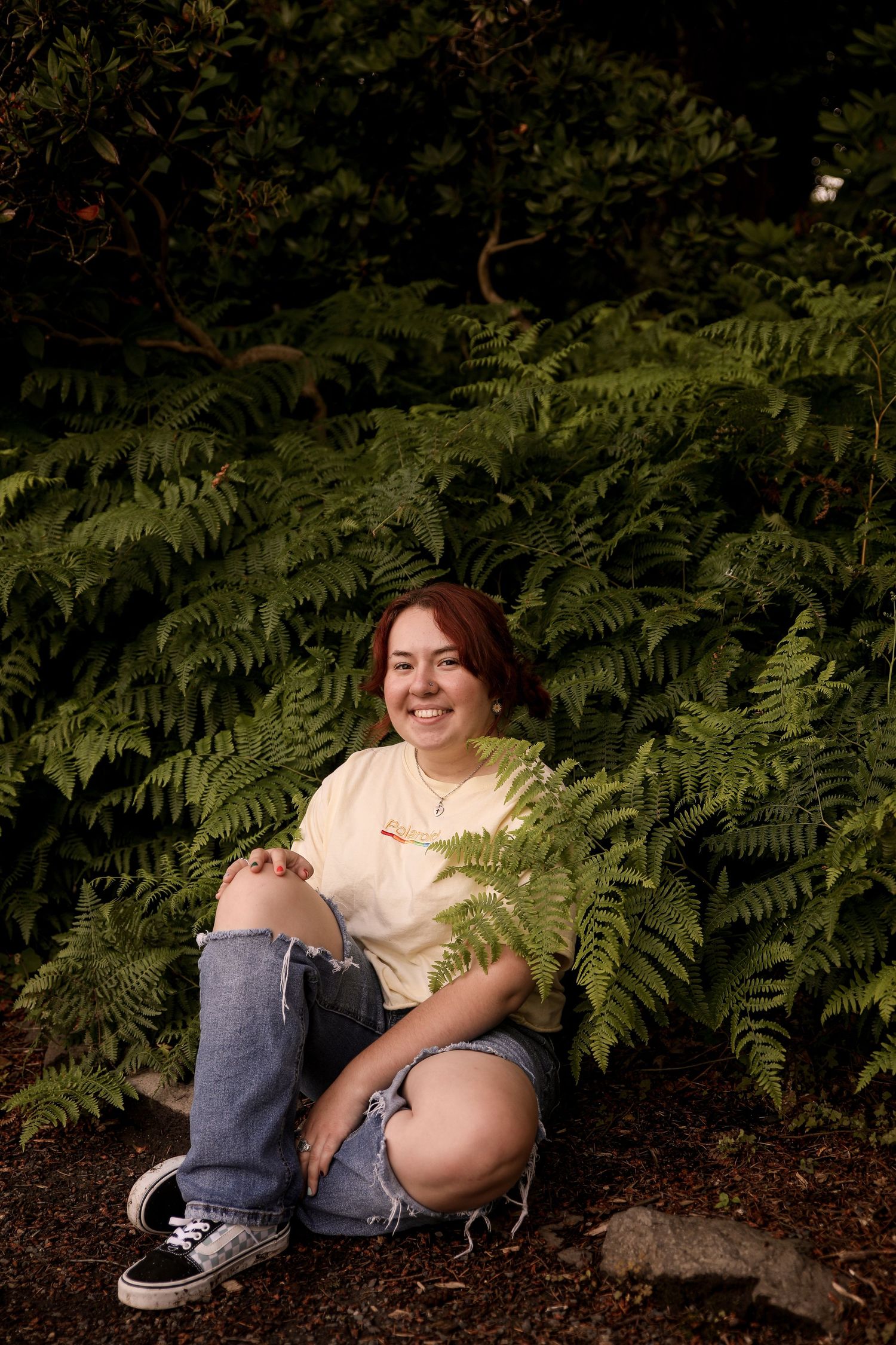 Person in casual attire sits by lush ferns in natural outdoor setting.