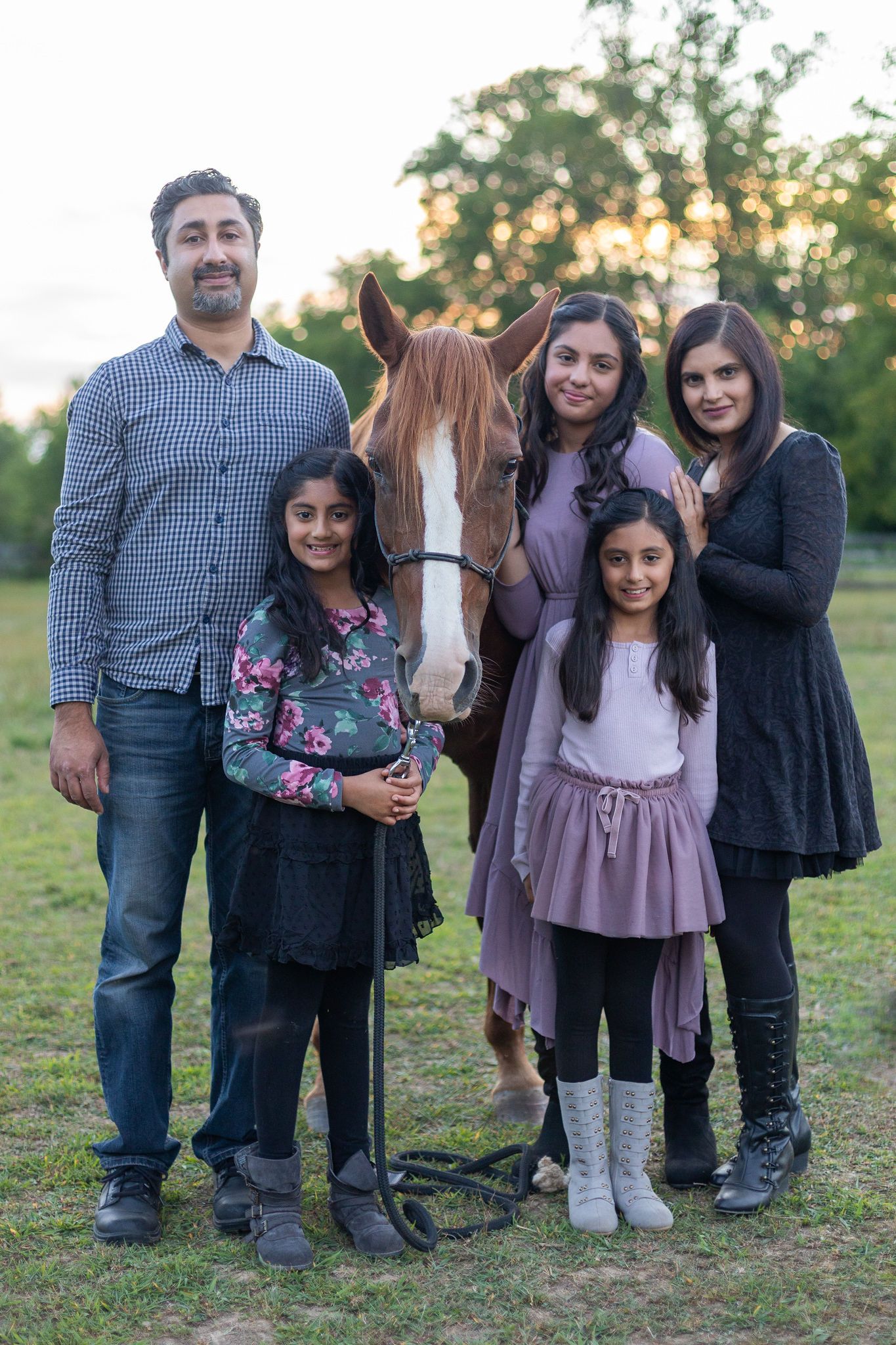 Family group stands beside their chestnut horse in a grassy field during sunset.