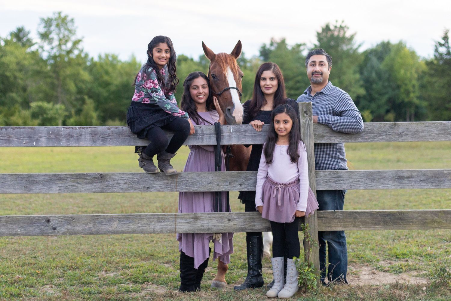 Family poses together by wooden fence with their brown horse in countryside setting.