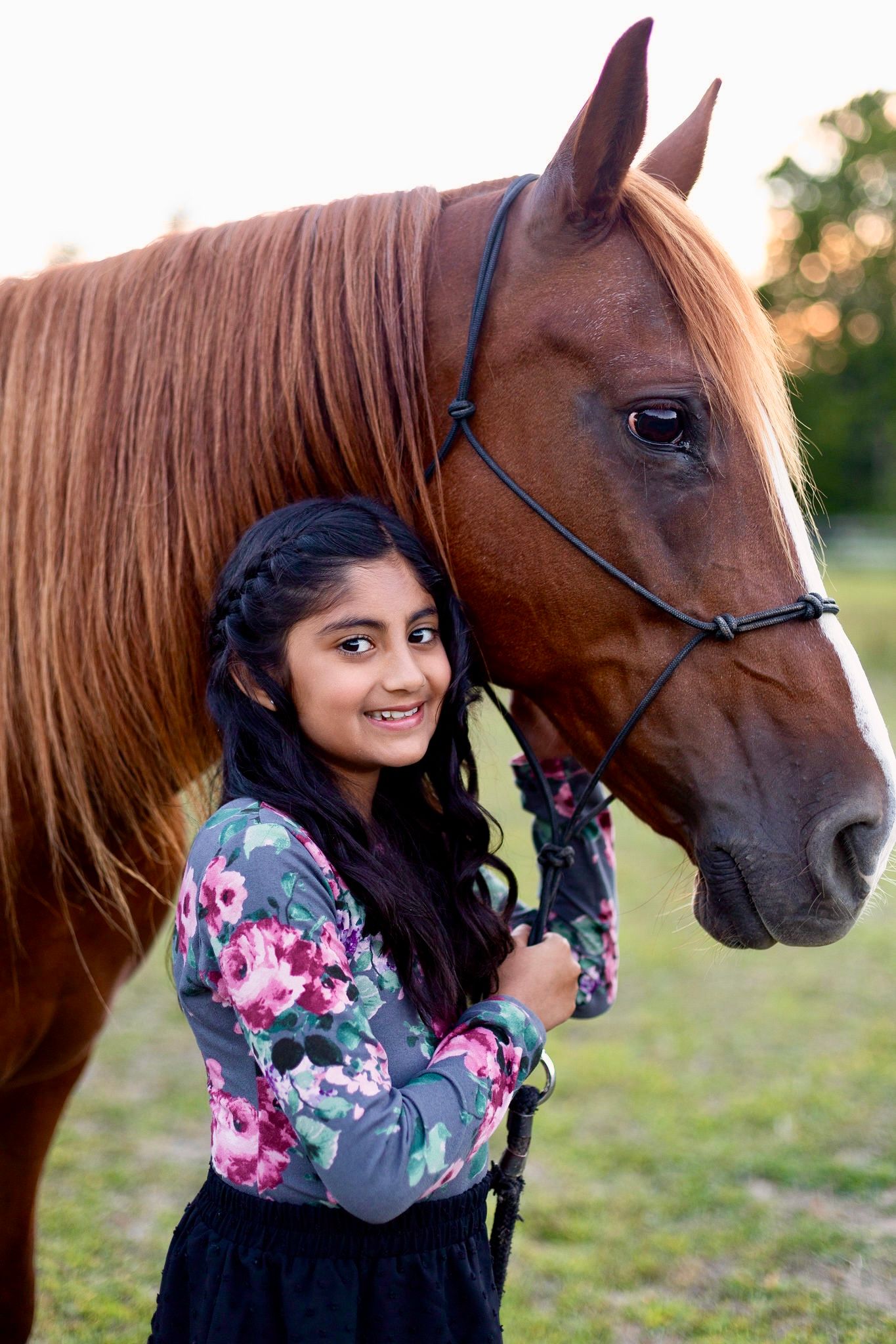 In a golden sunset, a person in a floral top stands next to a chestnut horse in a grassy field.