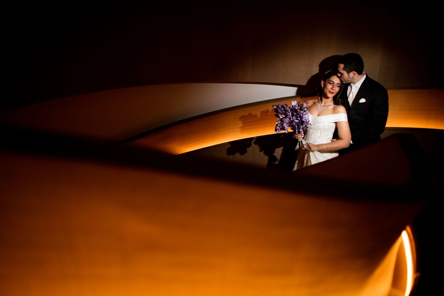 A romantic couple in formal wedding attire embrace in dramatic warm lighting for an artistic portrait.