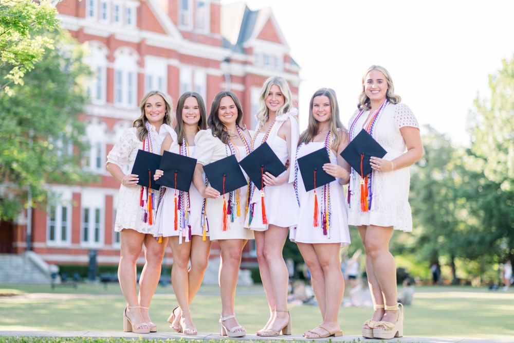 Group of graduates in white dresses pose together on university lawn with historic building backdrop.