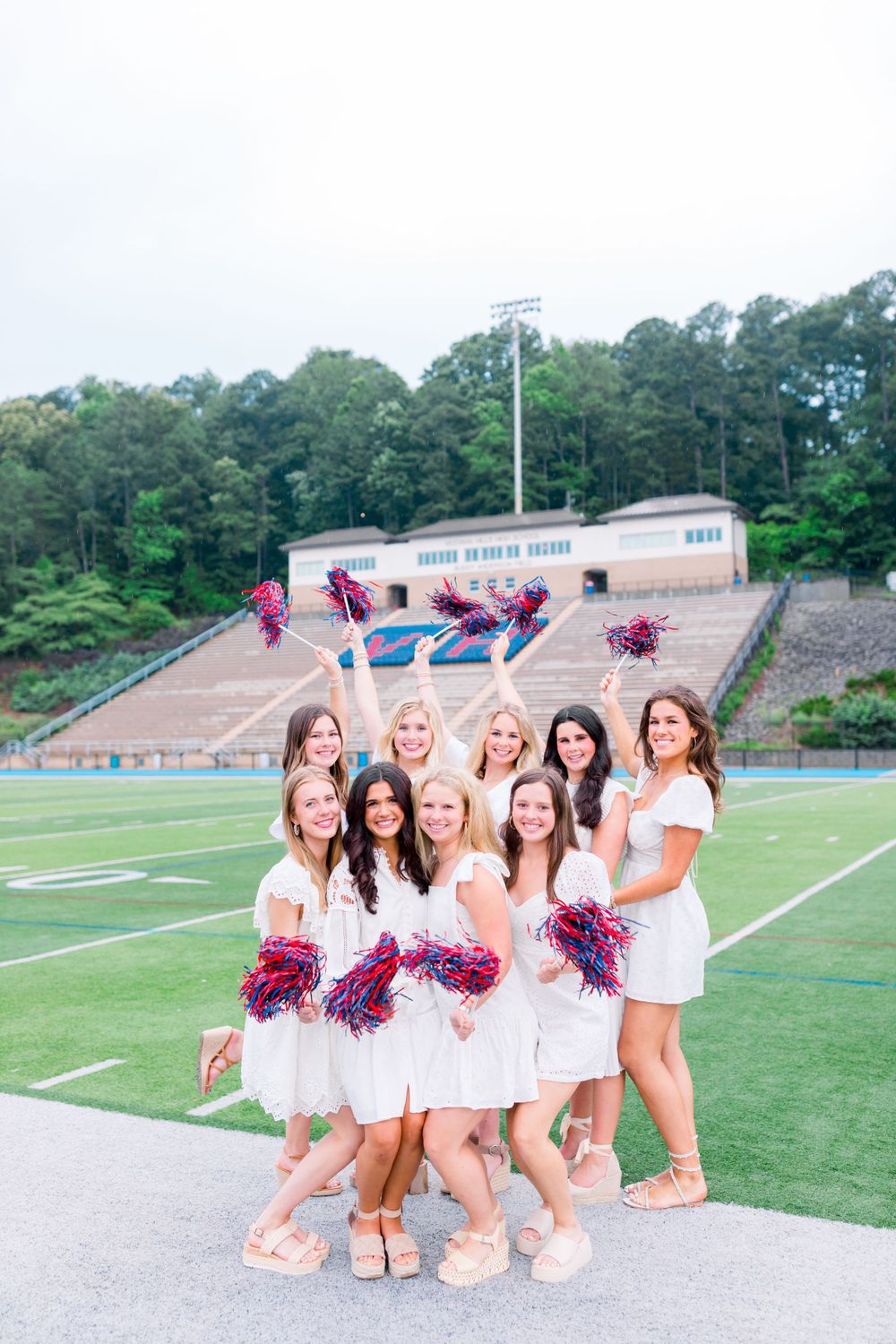 Cheerleaders in white outfits with red pom poms pose together on a football field.