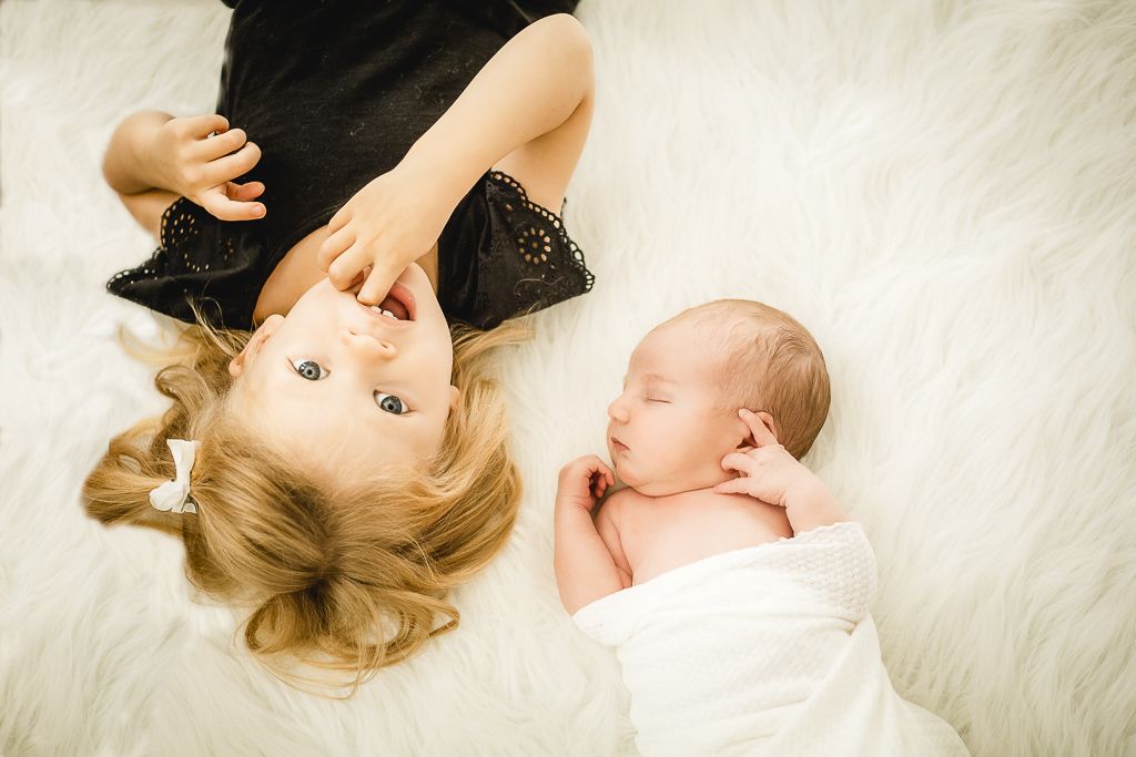 Two children lying on a white fur blanket, one sleeping while wearing a white wrap, and one in black attire.
