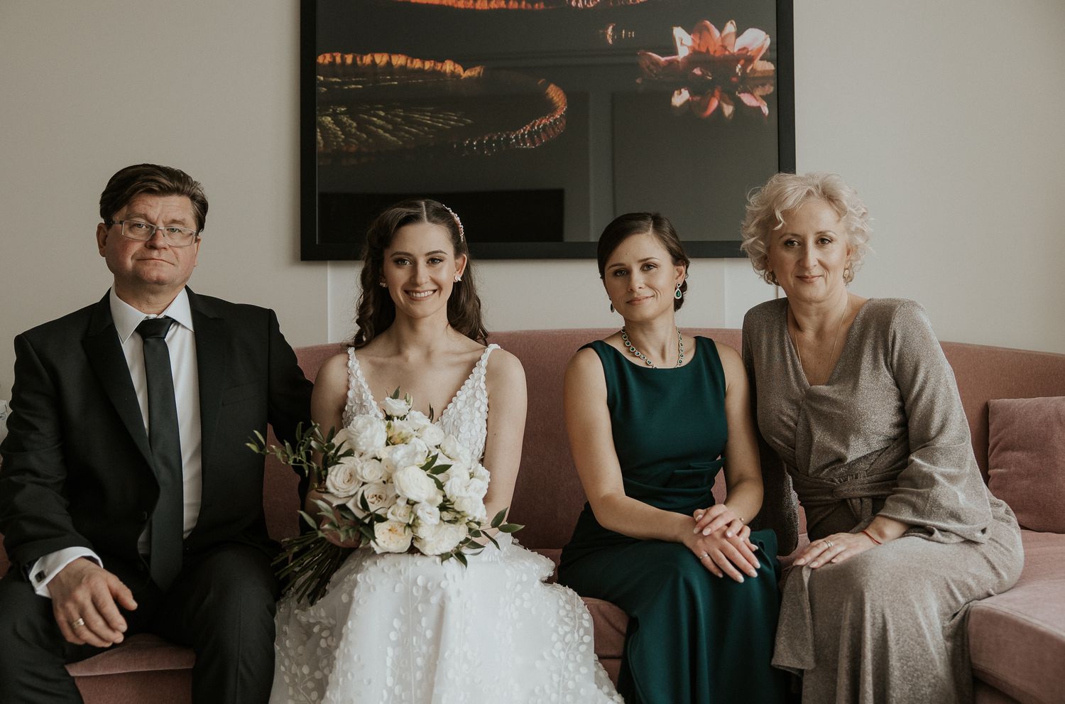 Bride with white bouquet poses with family on pink sofa before wedding ceremony.