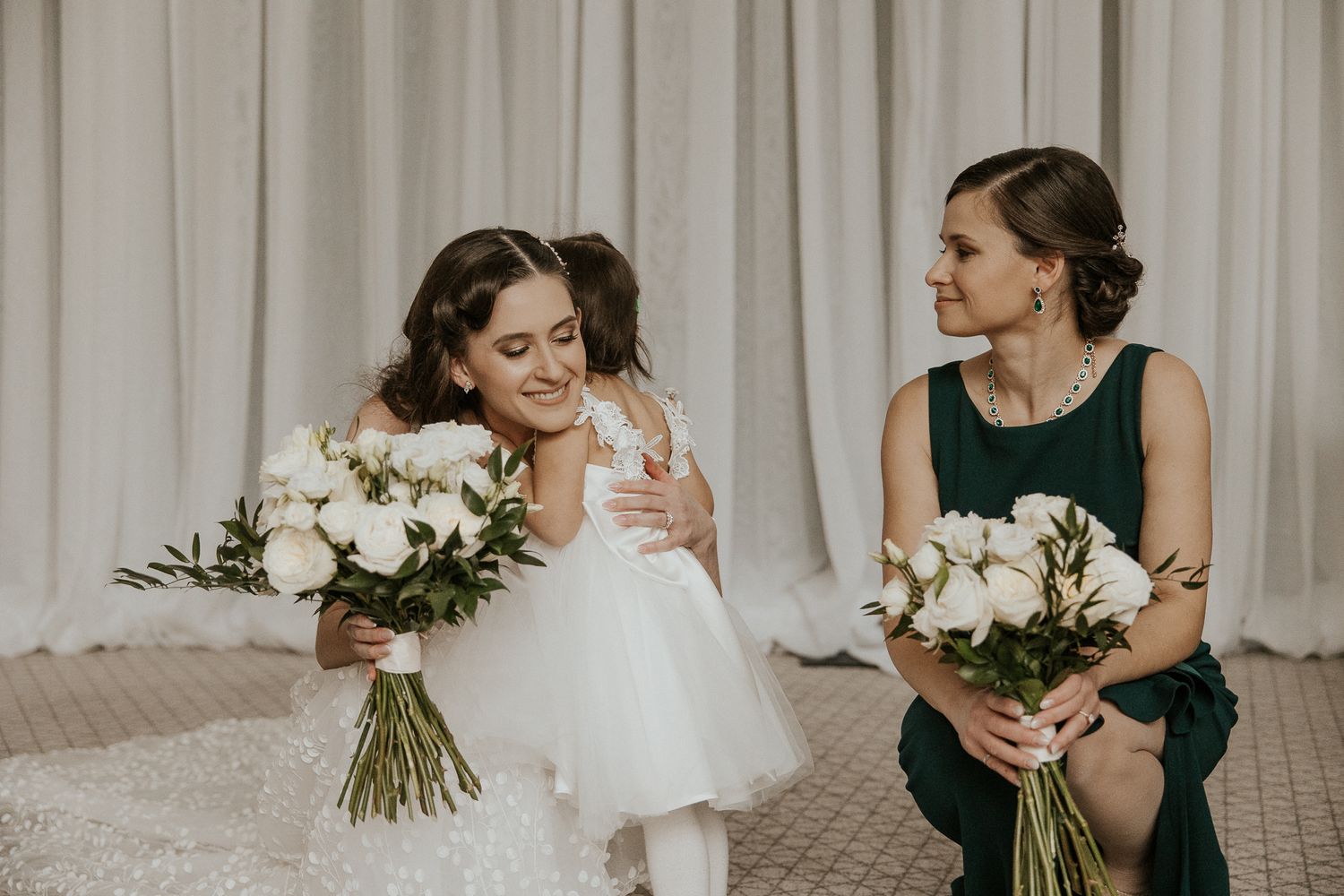 Bride kneels with flower girl both holding white rose bouquets in elegant bridal suite.