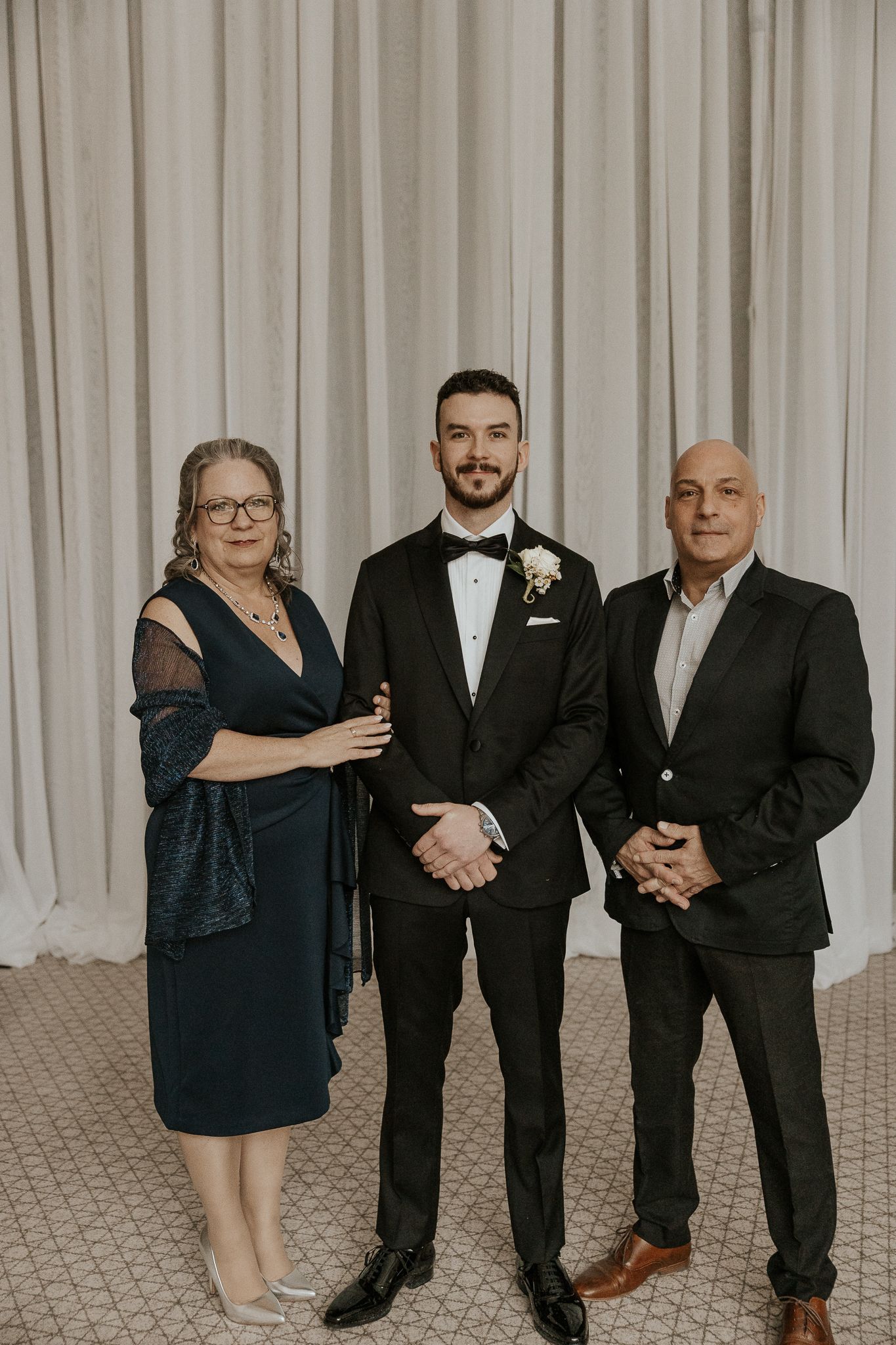 Groom in black tuxedo poses with parents in front of white draped curtain backdrop.