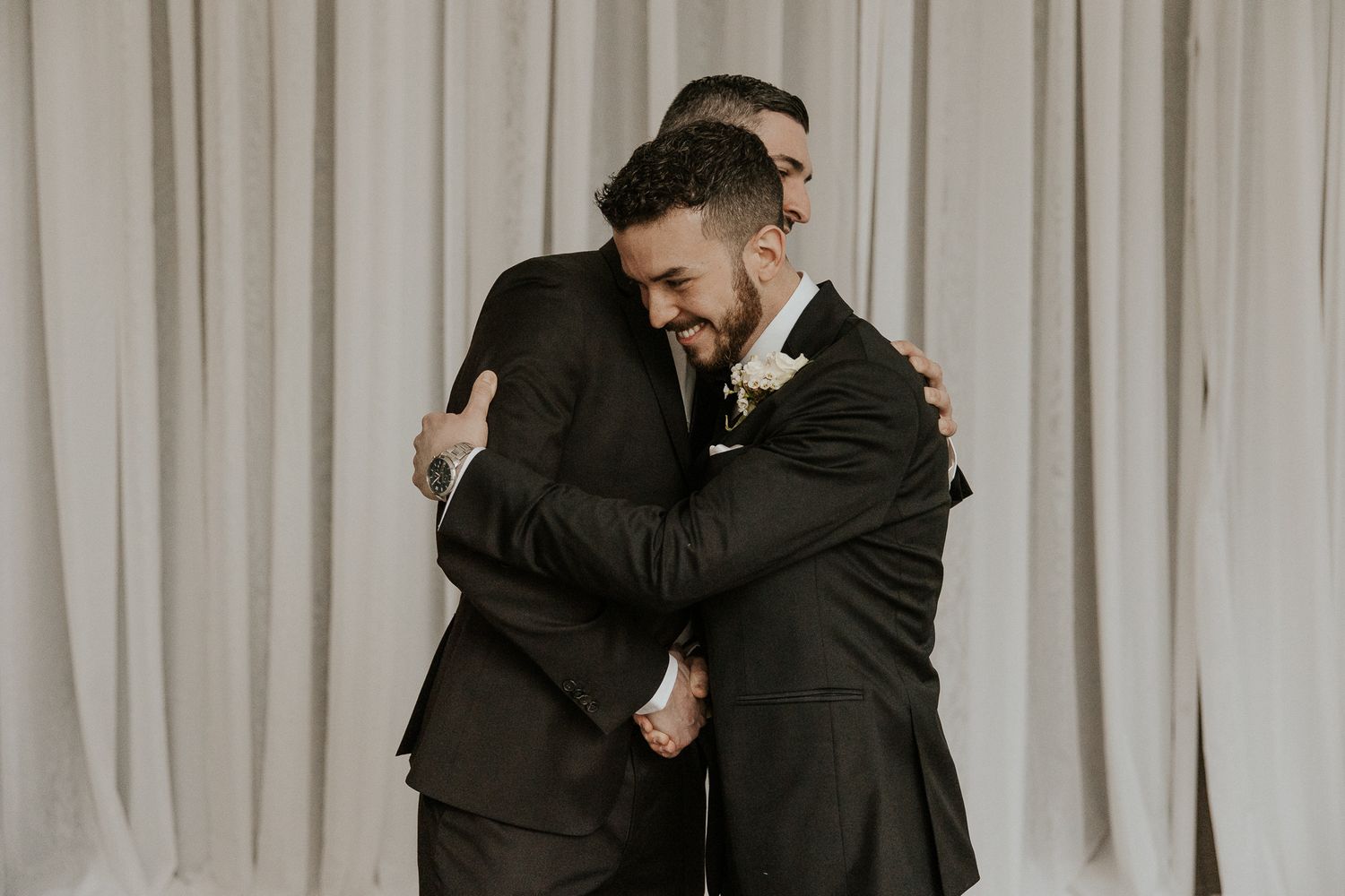 Groom shares warm embrace with groomsman in black suit at wedding venue.