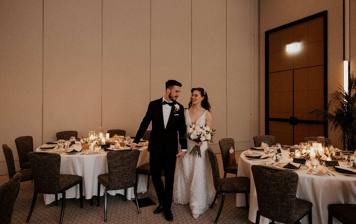 Newlyweds walk through candlelit wedding reception hall with elegantly set round tables.