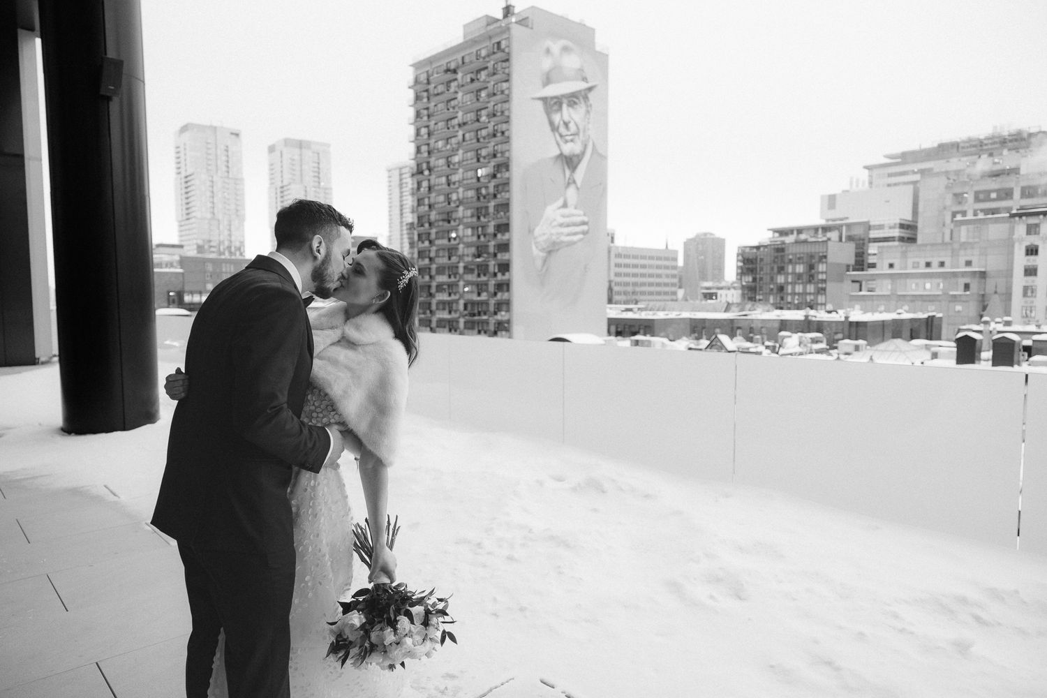 Bride and groom share romantic kiss on snowy rooftop with city mural backdrop.